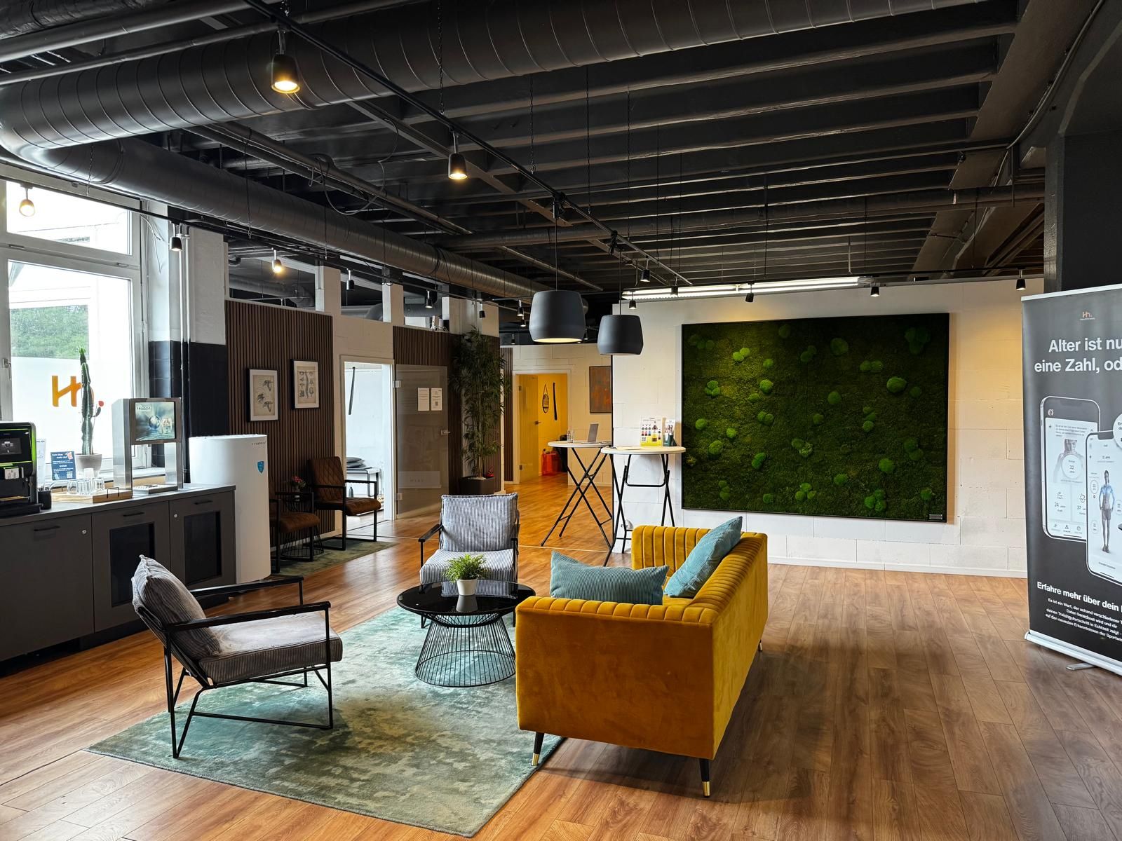 Die offene Lounge vor den Behandlungsräumen: ein senfgelbes Sofa vor einer grünen Mooswand, darüber eine offene Industriedecke
