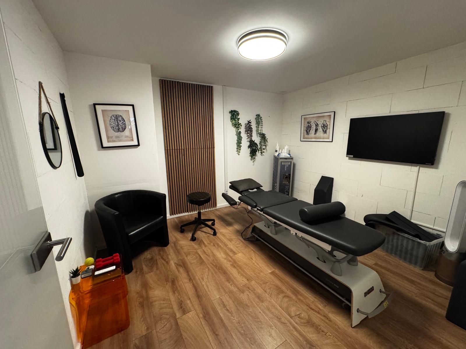 Blick von der Ecke in den Behandlungsraum: Liege mit Nackenrolle, Hocker, ein Bildschirm an der Wand und anatomische Drucke